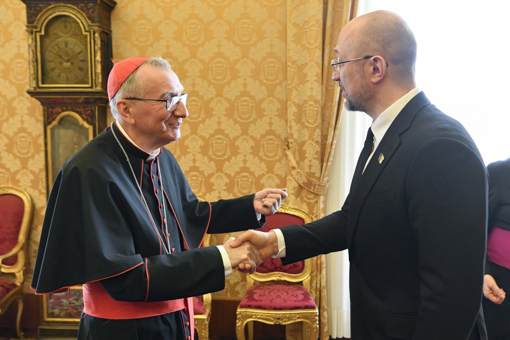 A man wears a cardinal's cassock and zucchetto and shakes a bald man in a suit's hand