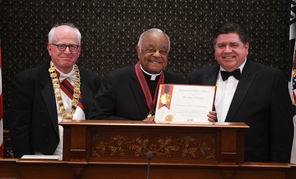 Cardinal Wilton Gregory, wearing a cassock and medal, holds a certificate and stands between two other men