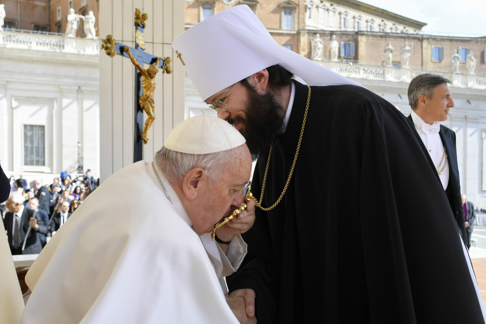 Pope Francis, sitting, leans forward to kiss something hanging by a chain around a man's neck, who wears a black robe and a white koukoulion
