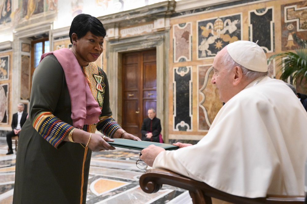 A Black woman who wears a olive green and multi-colored dress hands a folder to a seated Pope Francis