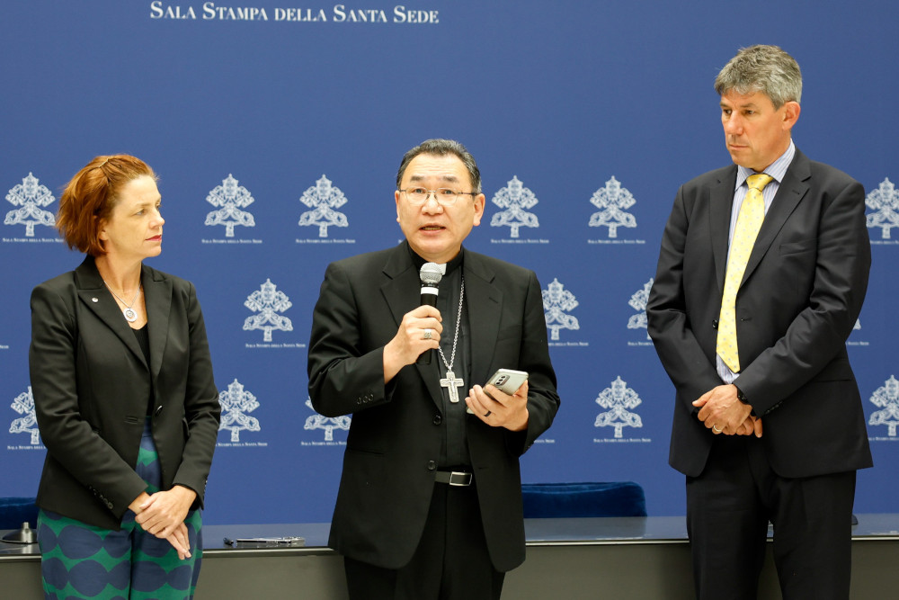 A man wearing a black suit, clerical collar, pectoral collar and glasses speaks into a microphone while a man and women in professional clothes stand on either side