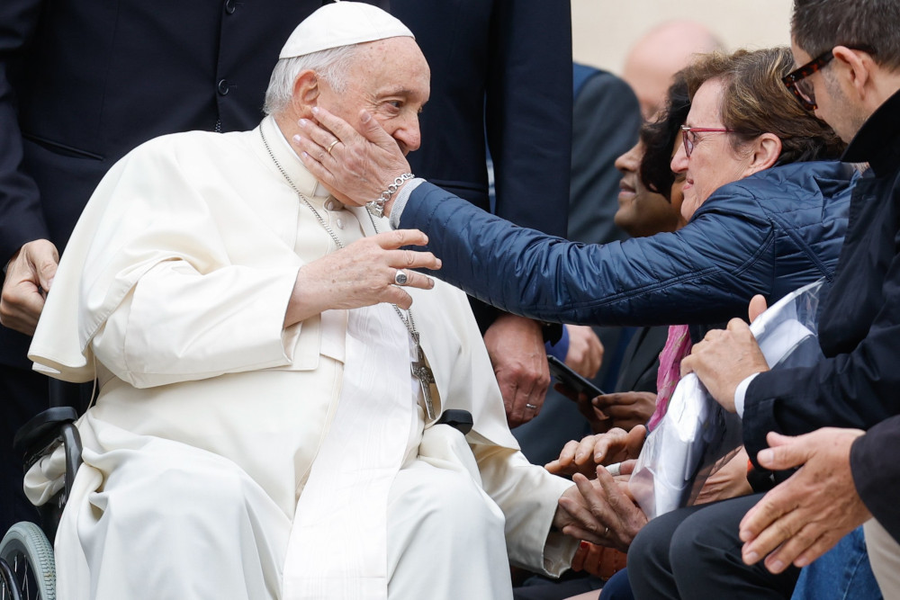 A person touches Pope Francis' cheek as he rides in his wheelchair past a line of people 