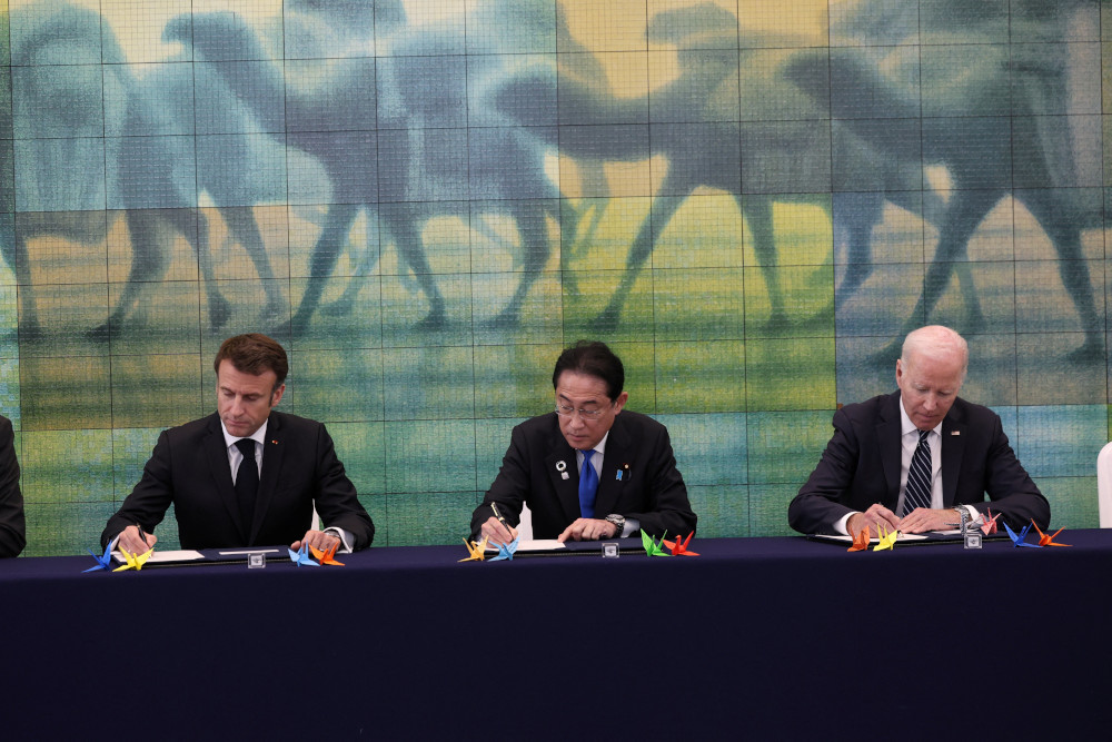 Three men in dark suits sit at a table with paper cranes on it below a painting of unidentifiable four-legged animal forms