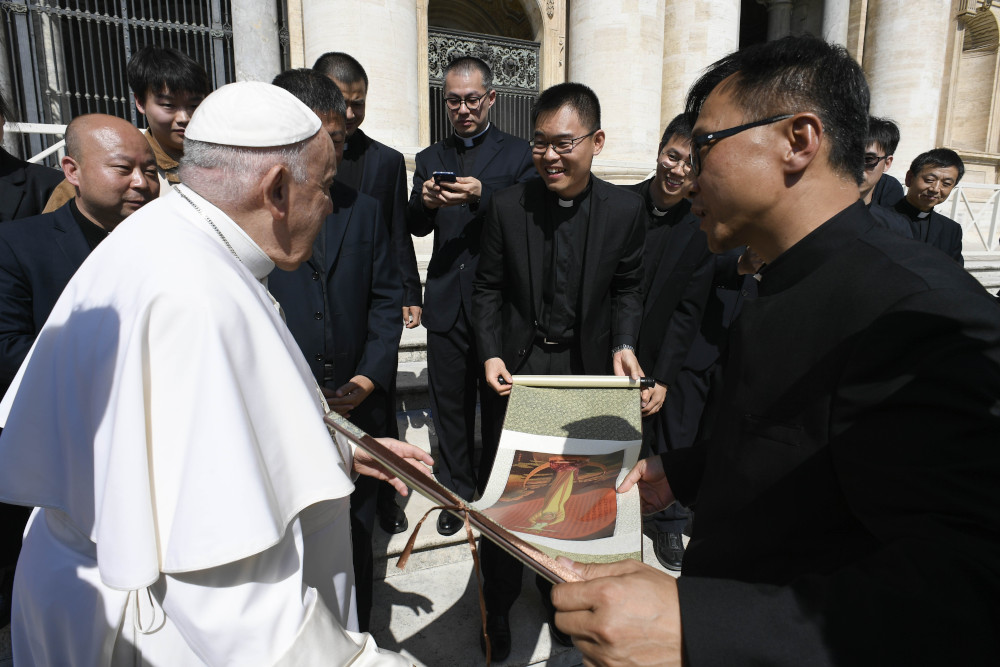 Pope Francis talks to a group of Chinese men wearing clergy collars and black suits. The men hold a unfurled scroll with artwork.