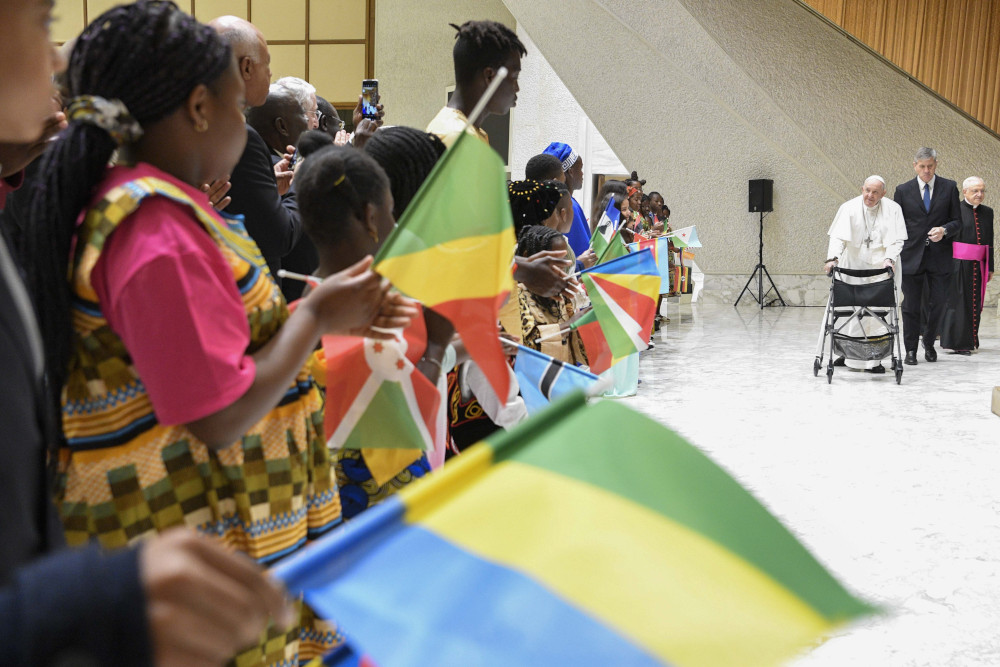 Pope Francis uses a walker to walk towards a long line of African children holding various colorful flags