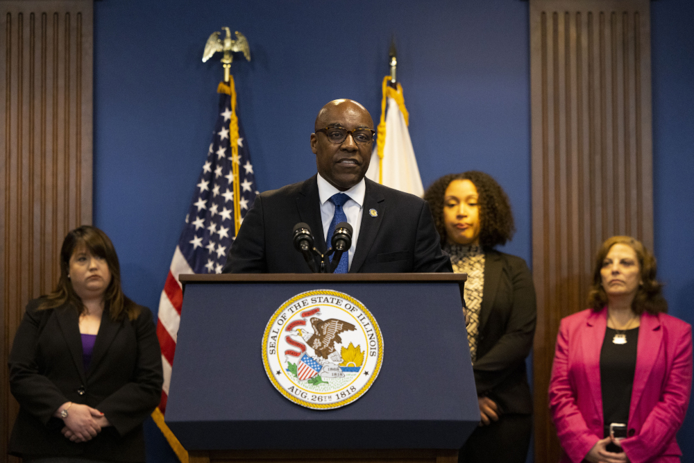 A bald Black man speaks behind a podium with an Illinois seal into several microphones, as flags and others stand behind him