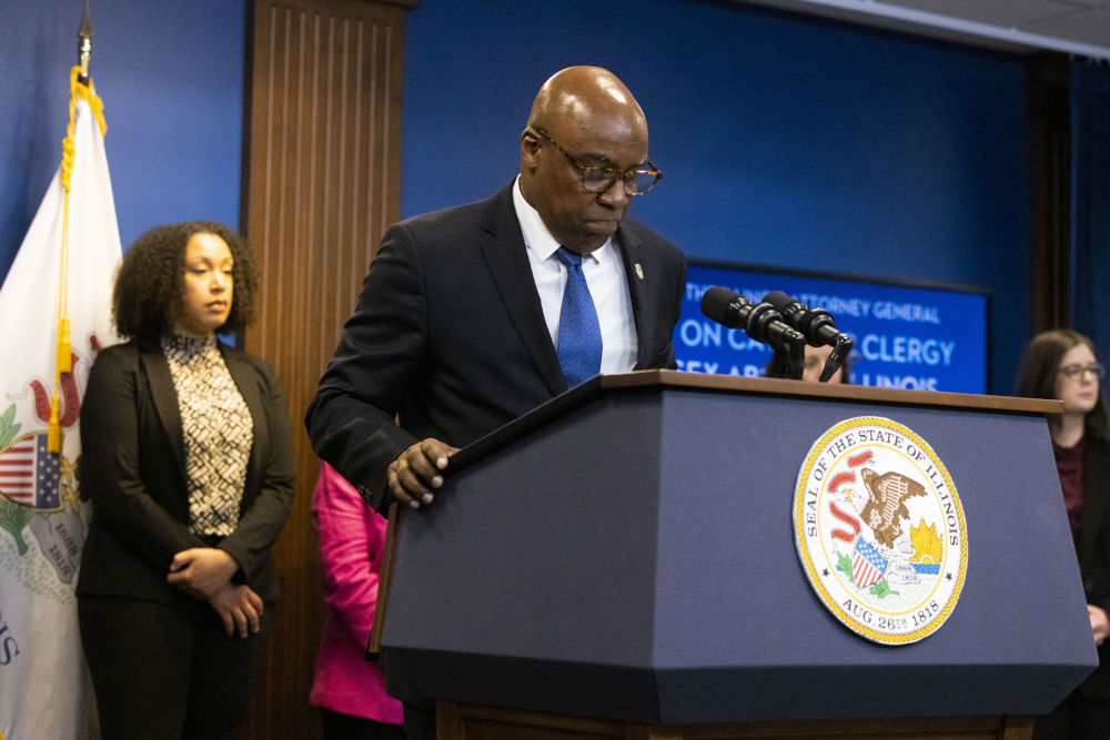 a Black bald man with glasses and a suit looks down at a podium with microphones