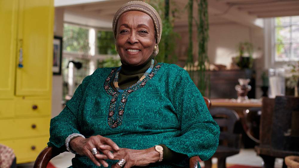 An older well-dressed Black woman wearing a head covering sits in a chair and smiles at the camera