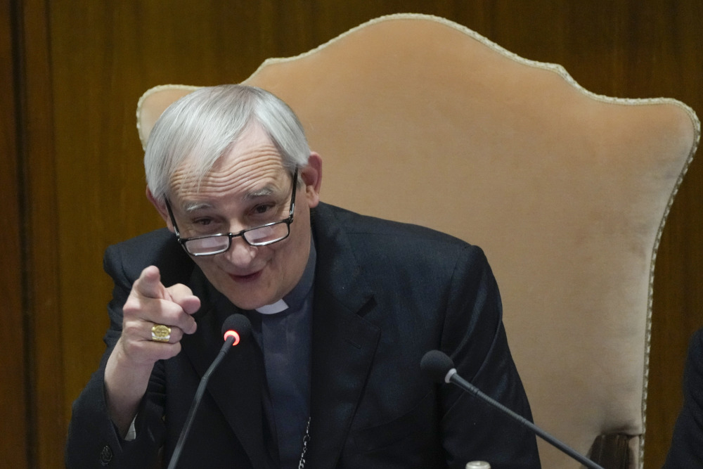 A man in a clerical collar and jacket speaks into a microphone and points his finger