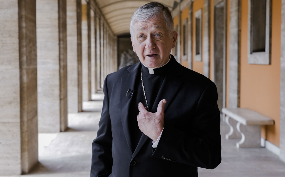 An older white man with blue eyes wears a clerical collar, black coat, pectoral cross, and microphone as he speaks to the camera