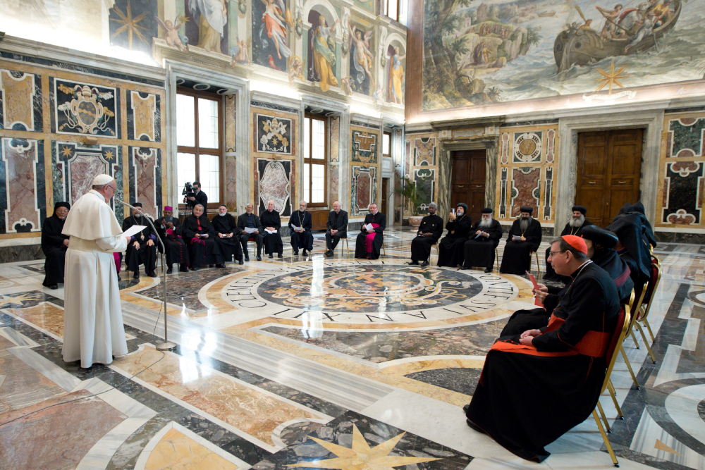 Pope Francis stands up and addresses a group of leaders in Roman Catholic and Orthodox garb in a room with Baroque art