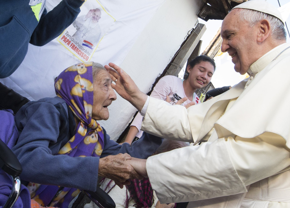 Pope Francis holds the hand of an older woman whose head is wrapped in a scarf as he blesses her with his other hand