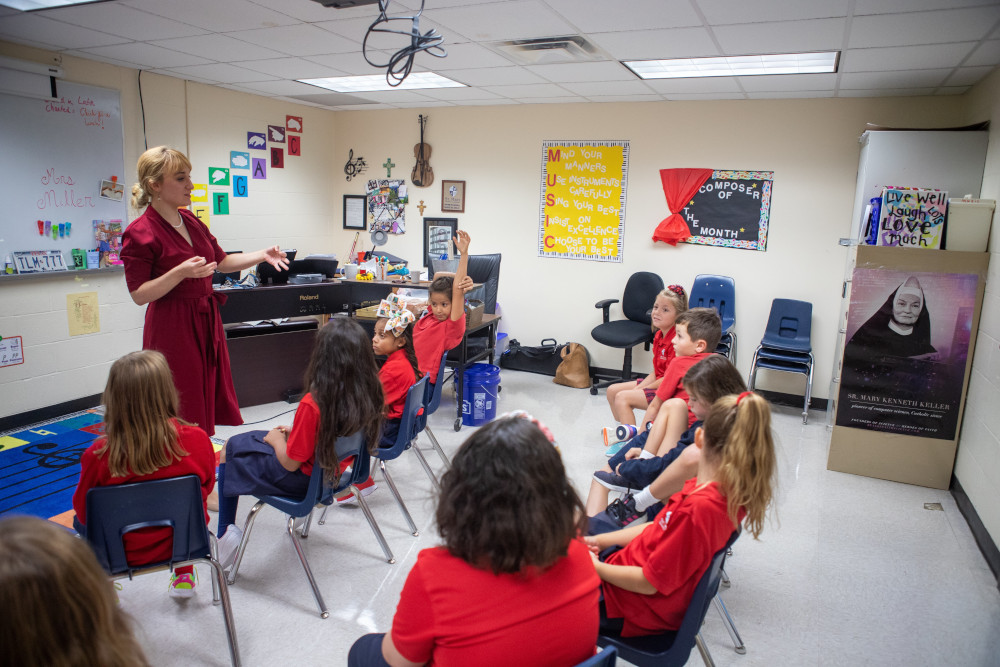 A blonde woman in a red dress stands in front of a group of children in red uniforms in a music classroom.