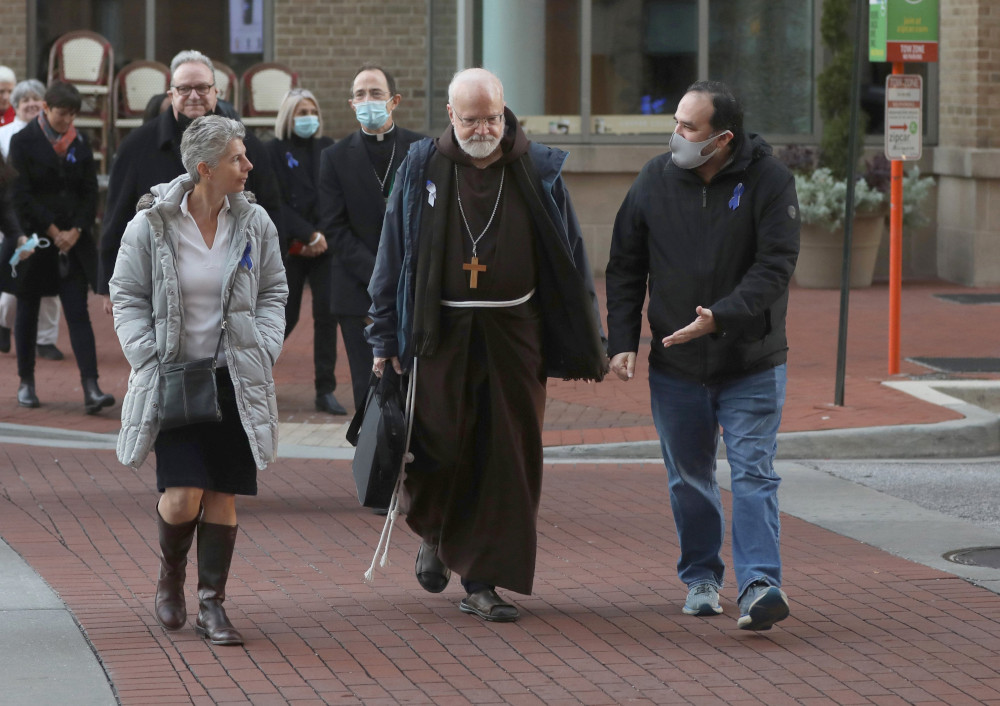 An older white man wearing a brown Franciscan habit walks between laity and priests. Some where face masks.