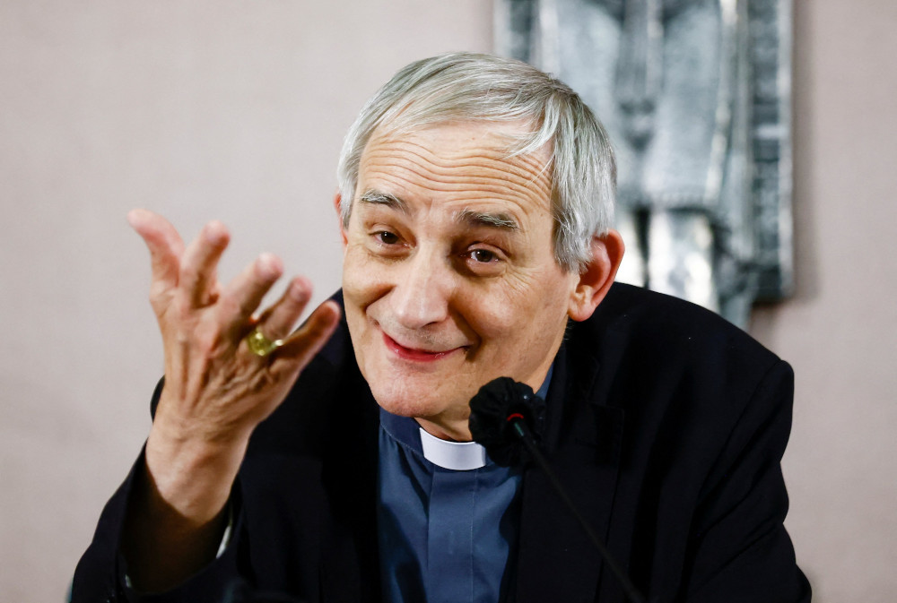 An older white man wearing a blue shirt, black jacket and clerical collar speaks into a microphone