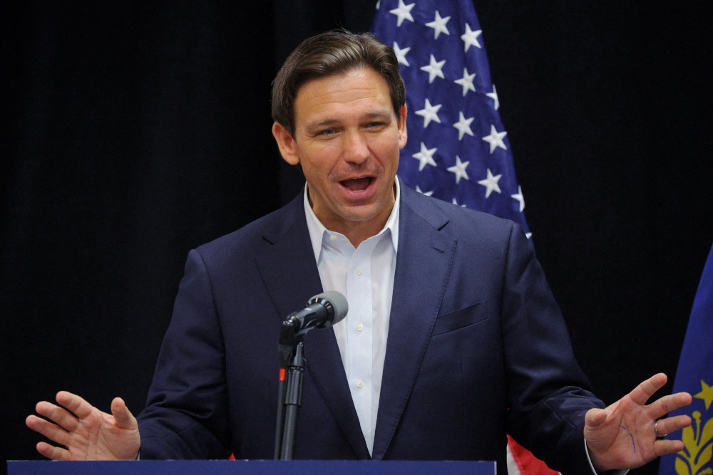 Ron Desantis wears a suit jacket with no tie and speaks into a microphone while standing in front of an American flag