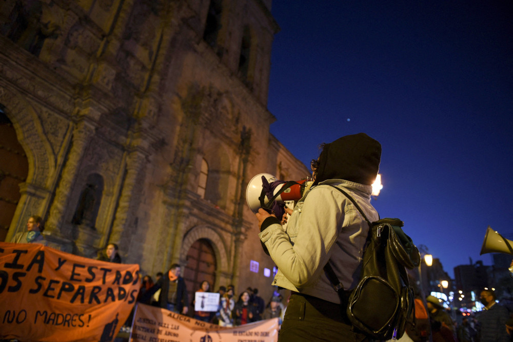 A person holds a megaphone while facing a cathedral at night. People with hand-painted banners stand in front of the cathedral