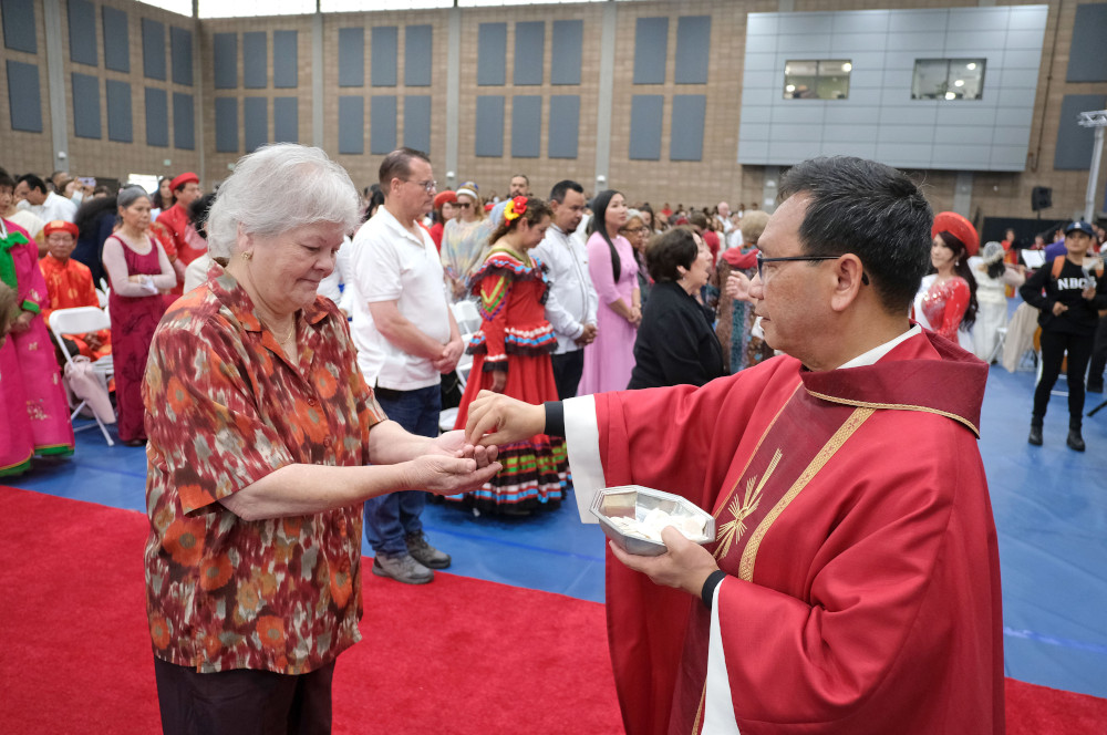 A black-haired man wearing glasses and a red chasuble places a Communion wafer in an older woman's hands