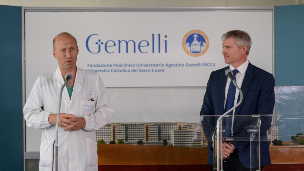 A white man in a white doctor's coat with scrubs peaking through stands next to a white man in a blue suit and tie. Both use microphones in front of a banner that says Gemelli