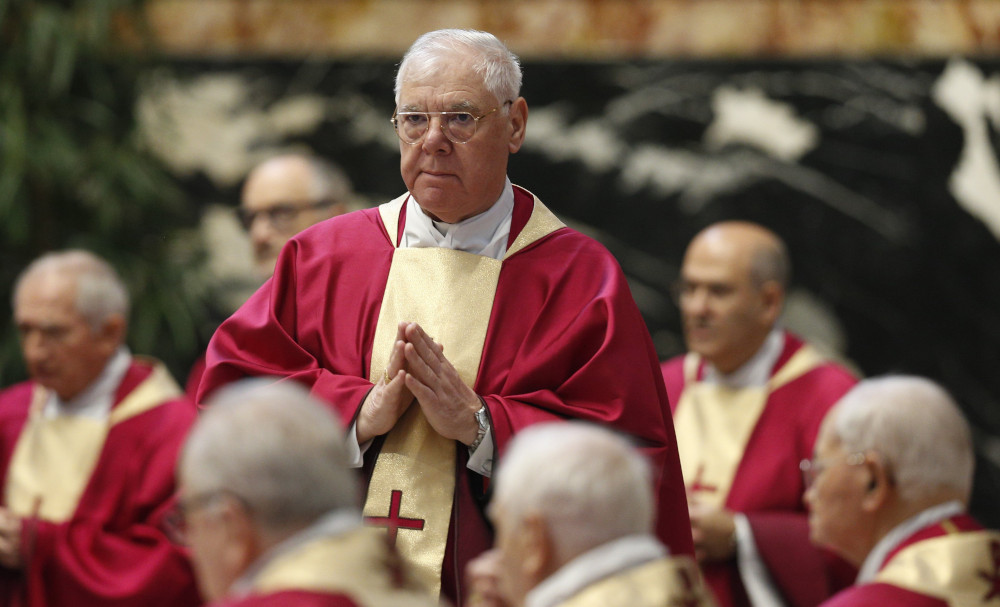 An older white man wears a red chasuble with a gold piece down the middle with a red cross on it. He stands with his hands together in front of him.