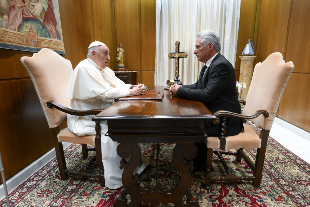 Pope Francis sits in a chair on one side of the table as an older white-appearing man sits in an identical chair on the opposite side