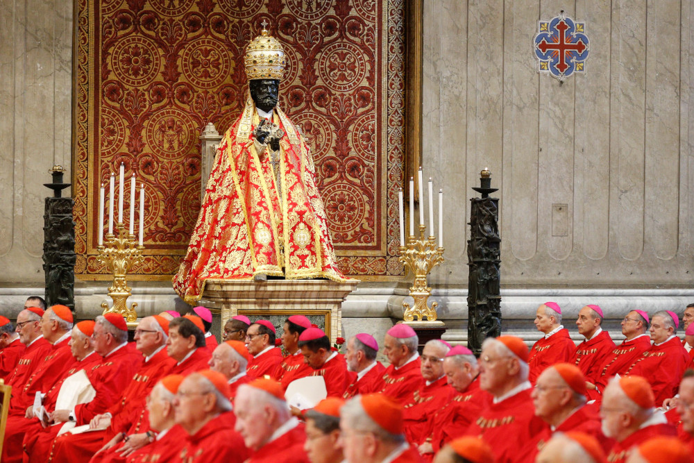 A black statue of St. Peter wears a red and gold robe and a red and gold dome-like tiara while towering over a large group of cardinals and bishops