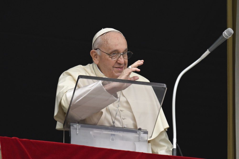 Pope Francis raises his hand to wave while standing behind a clear lectern and a microphone