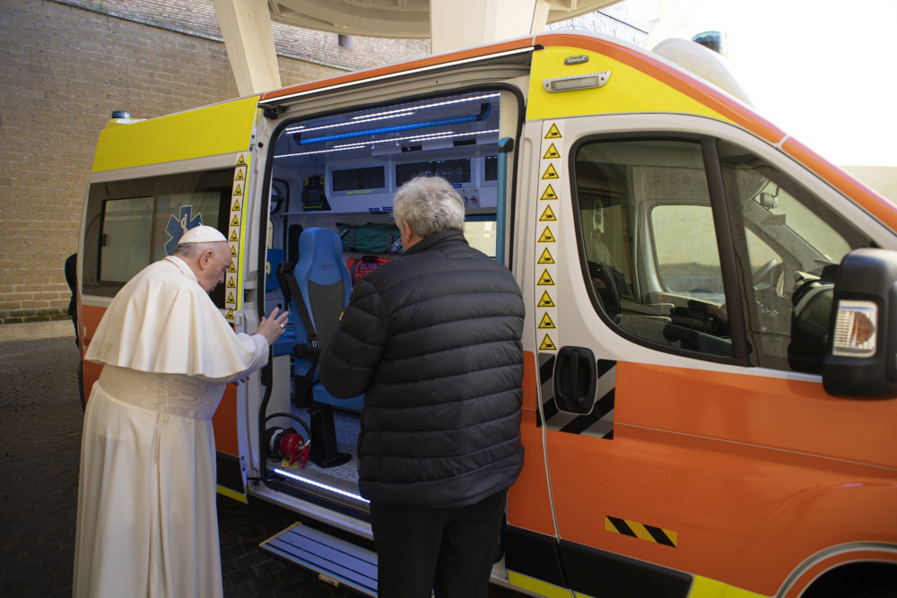 Pope Francis raises his hand as he stands outside an orange and yellow vehicle next to a man with white hair