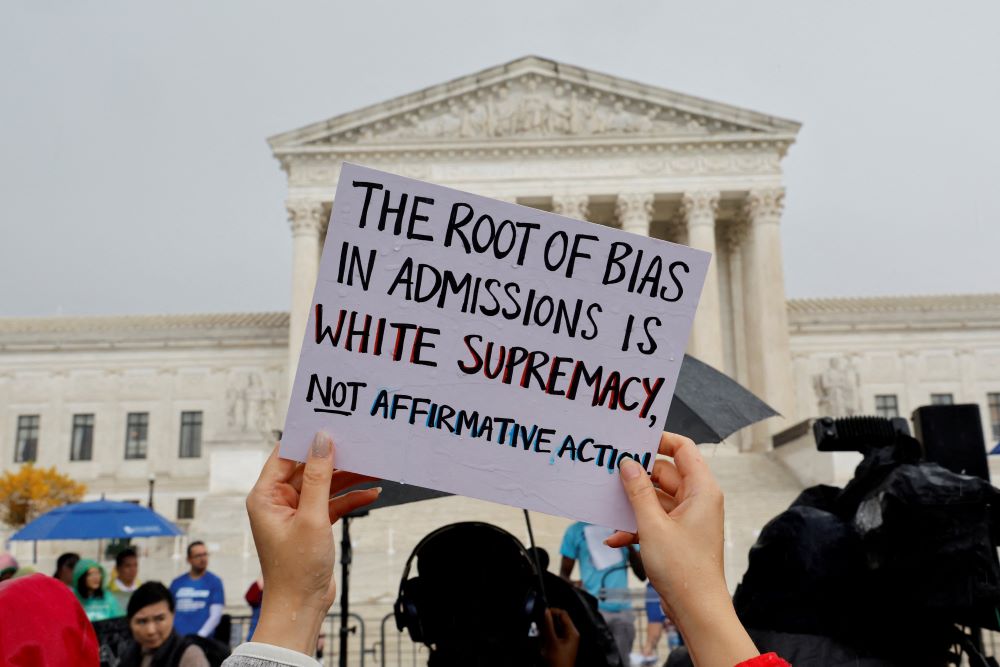 Demonstrator holds sign outside Supreme Court. It reads, "The root of bias in admissions is white supremacy, not affirmative action."