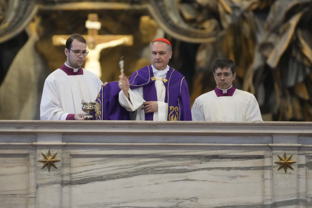A white man wearing a red zucchetto uses an aspergillum to sprinkle holy water on an alter. Two men in clerical collars stand beside him.