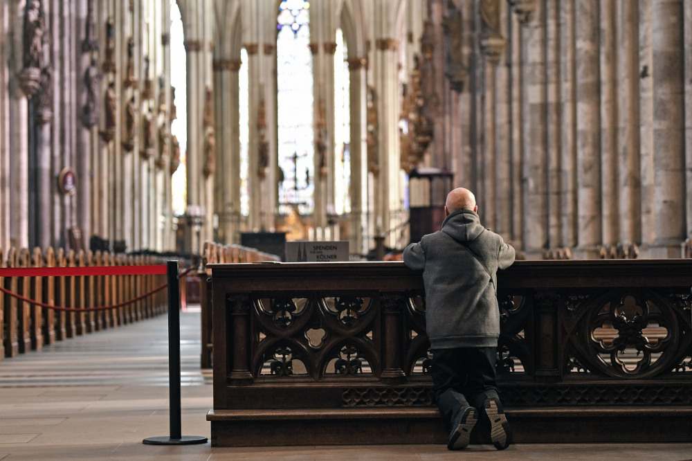 A man kneels on a kneeler in an empty-looking church with larger pillars lining the sides