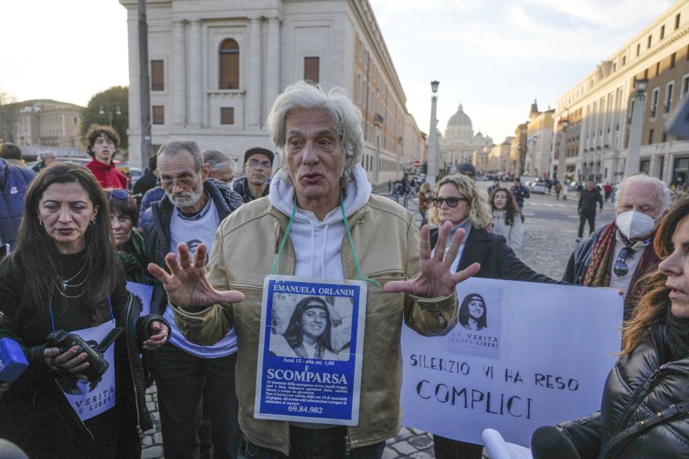 An older white man, surrounded by a crowd, talks with his hands and wears a sign around his neck with a picture of a girl on it