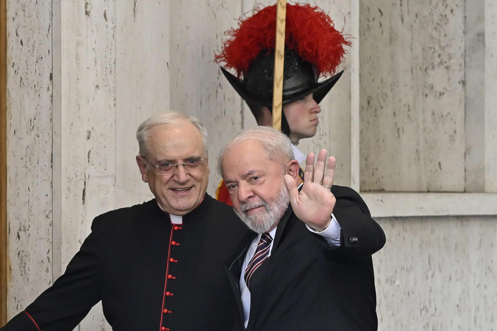 An older white man in a suit waves at the camera next to a man wearing a cardinal's cassock with a member of the Swiss Guard in the background