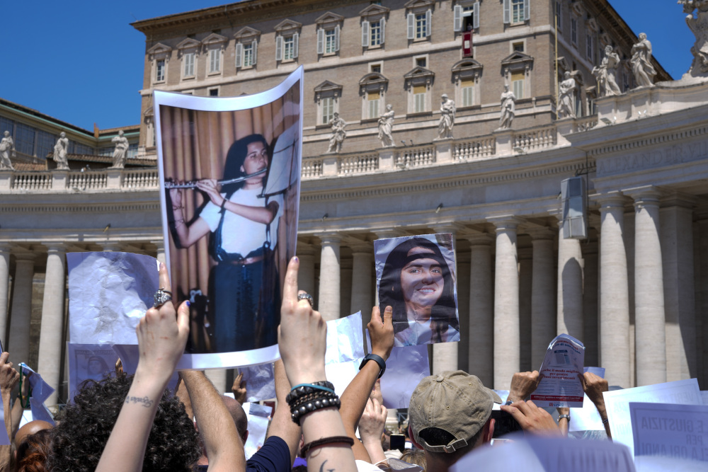 People hold up pictures of a black-haired girl, including one where she is playing the flute, while standing in St. Peter's Square below Pope Francis' open window