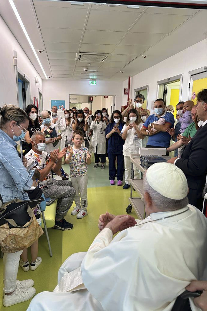 A large group of people, adults and children, many wearing masks or holding cellphones, stand around Pope Francis in a hospital hallway