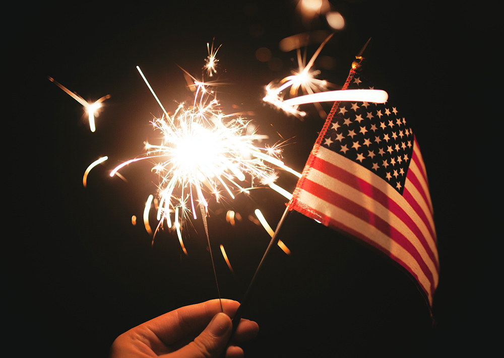 A hand holding a lit sparkler and a small American flag (Unsplash/Stephanie McCabe)
