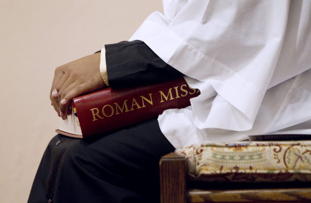 An altar server holds a red book with the gold letters "Roman Mis" visible.