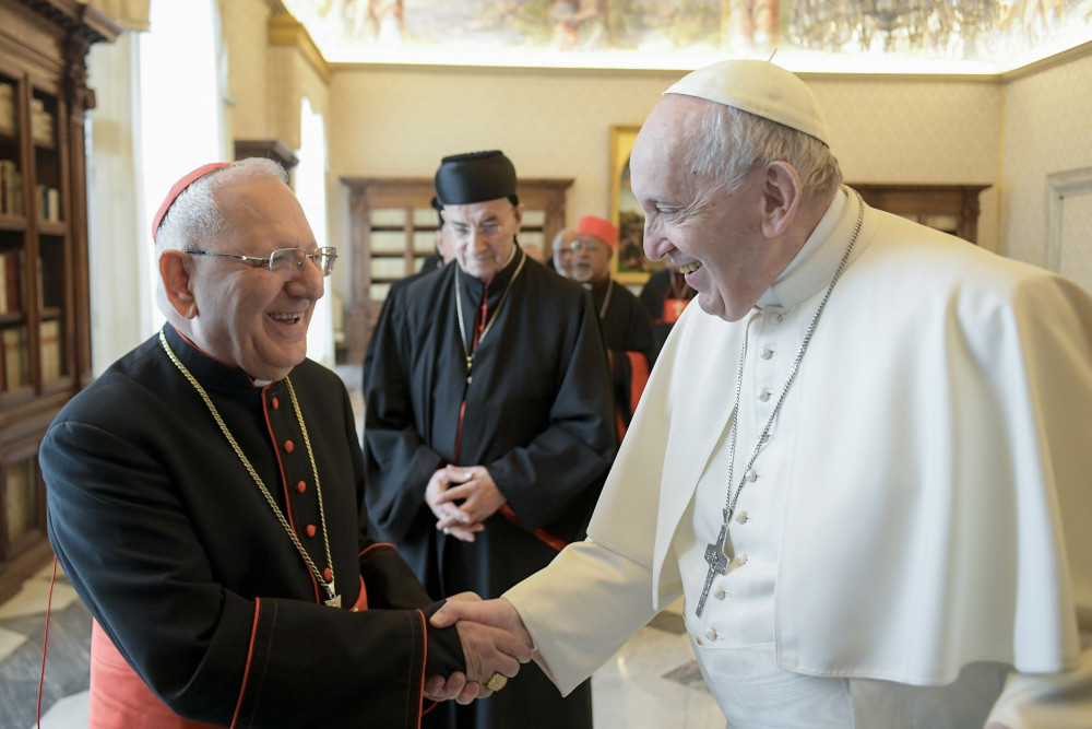 Pope Francis shakes the hand of a smiling white-haired man wearing a red zucchetto and a pectoral cross