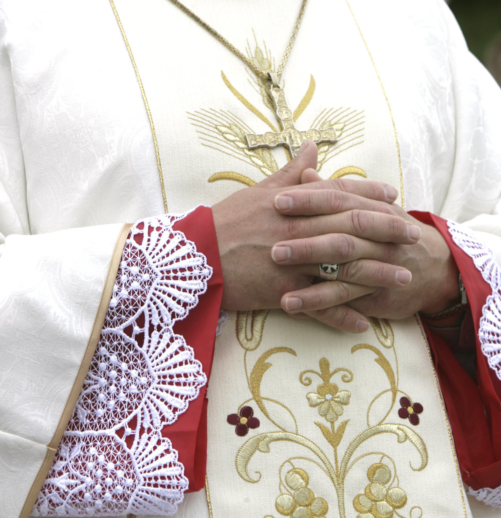 A white man's hands are folded as he stands in intricately embroidered white vestments. His head is not visible.