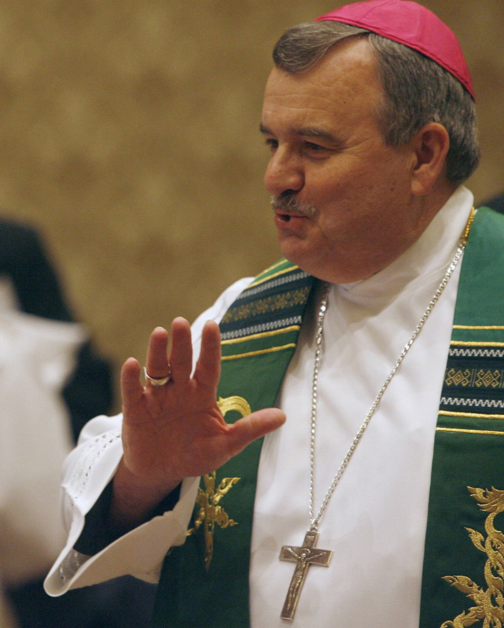 A white man with a moustache wears a violet zucchetto and green stole over white vestments as he talks and gestures