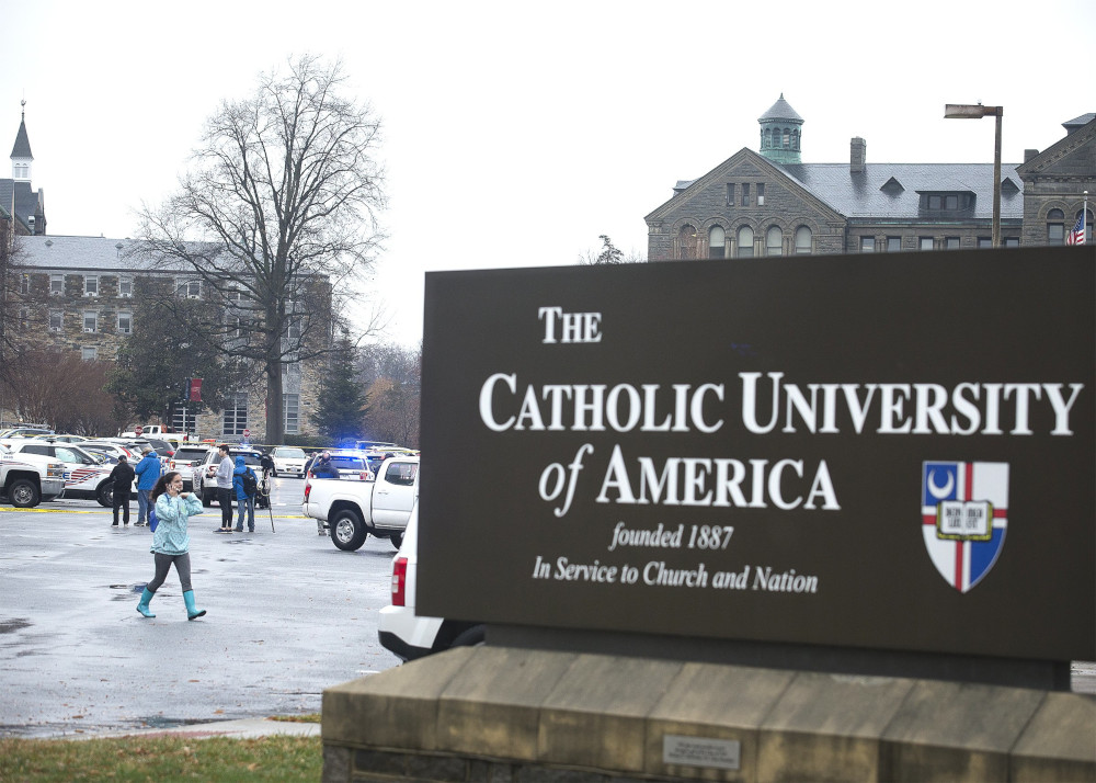 A sign that says "The Catholic University of America founded 1887 In Service to Church and Nation" is visible in front of a parking lot with police cars.