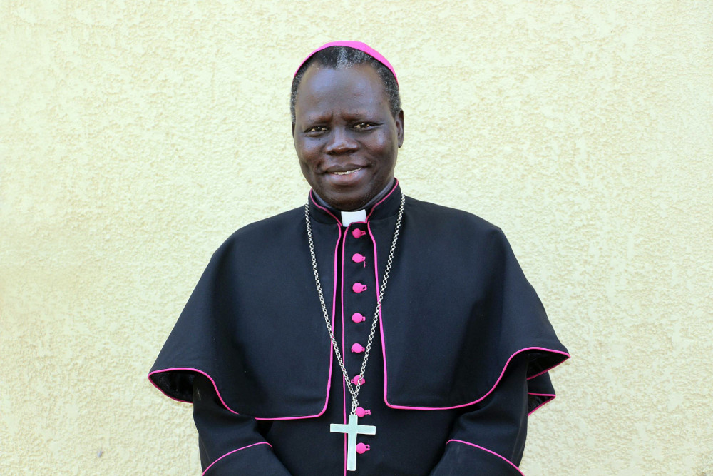 A Black man stands before a white wall and wears a violet zucchetto, pectoral cross and bishop's cape