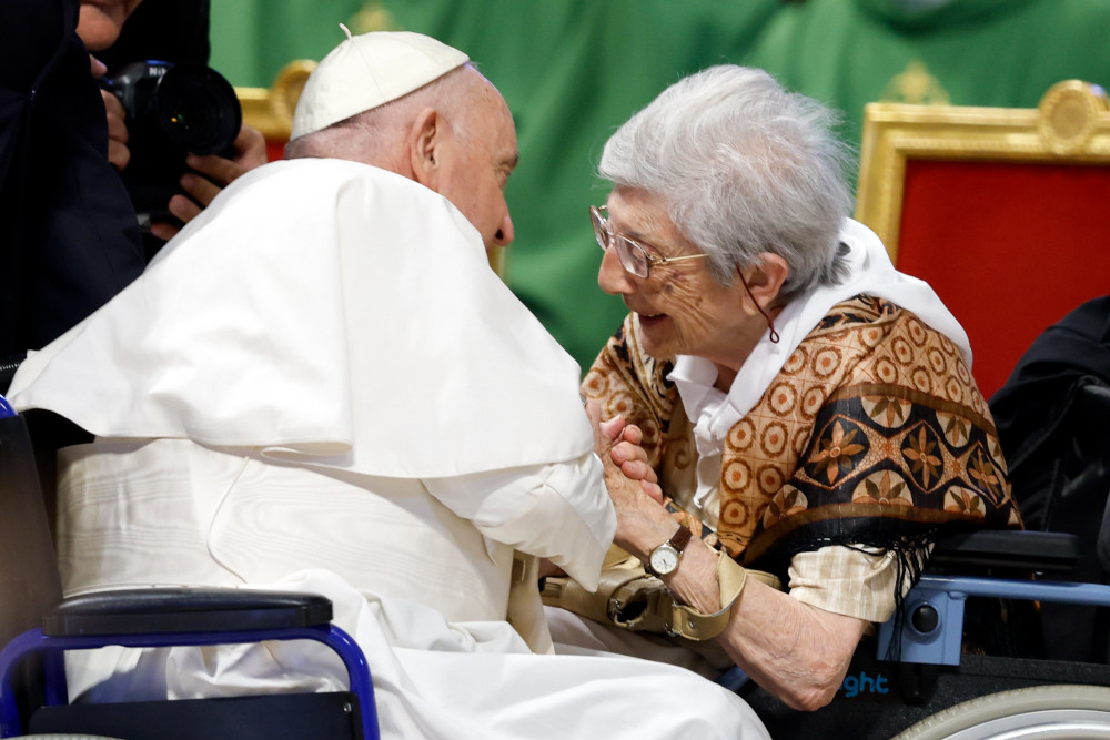 Pope Francis, seated in a wheelchair, leans forward to clasp the hands of an older white woman sitting in a wheelchair 