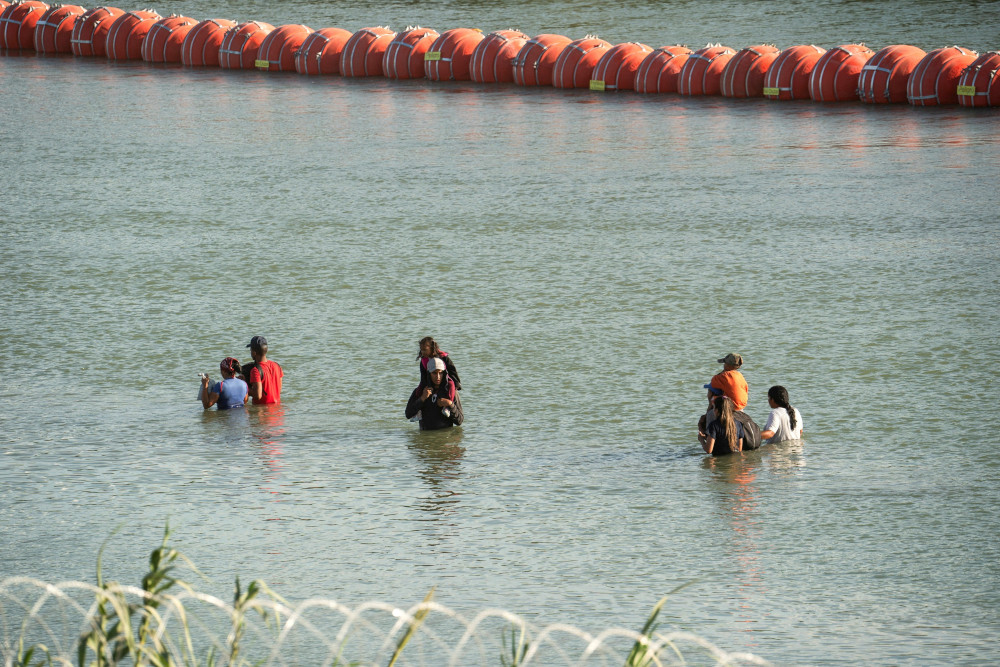 People stand in the middle of a river. Some have others on their shoulders. One part of the river is blocked by large orange buoys spaced so close as to be a wall and the other side is blocked by concertina wire.