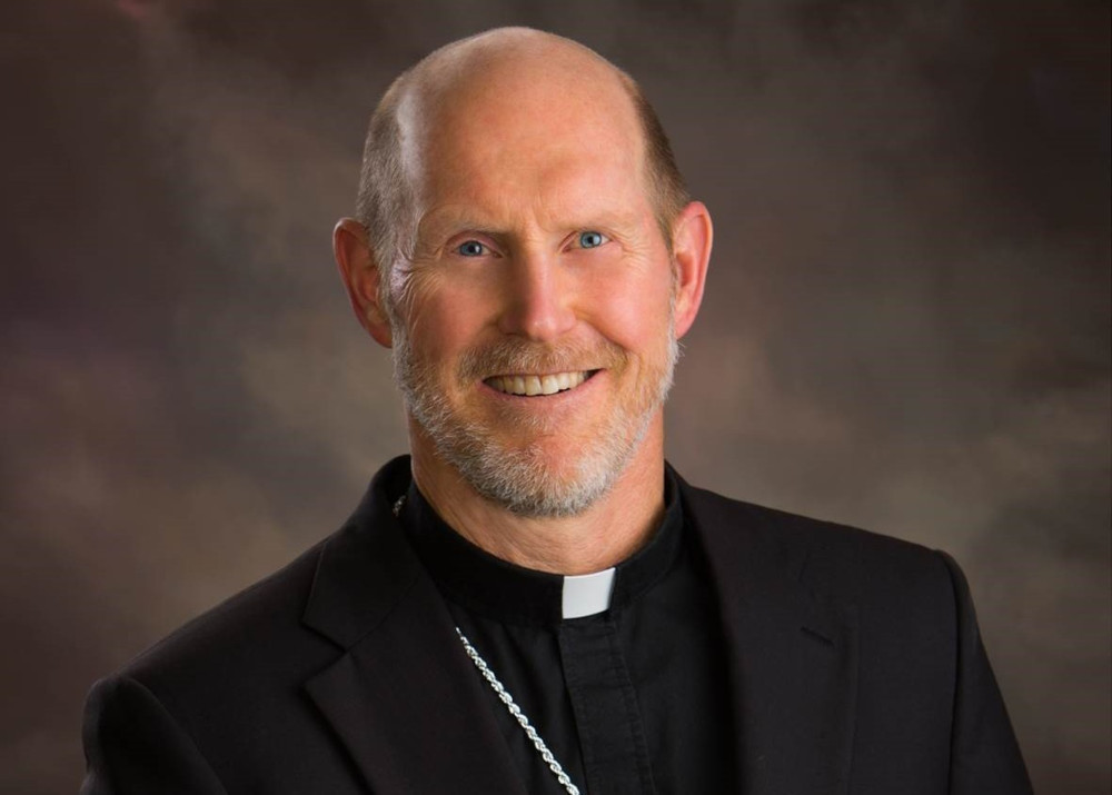 A white man with blue eyes and a beard wears a clerical collar and smiles at the camera