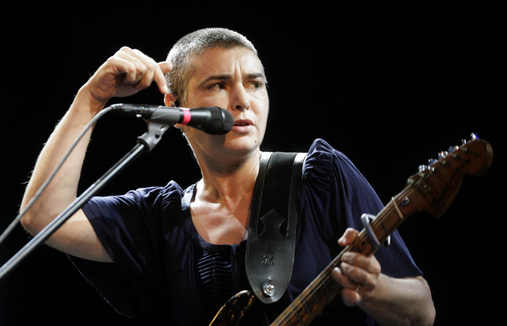 A white woman with a buzz cut holds a guitar and sings into a microphone while making a hand gesture pointing down