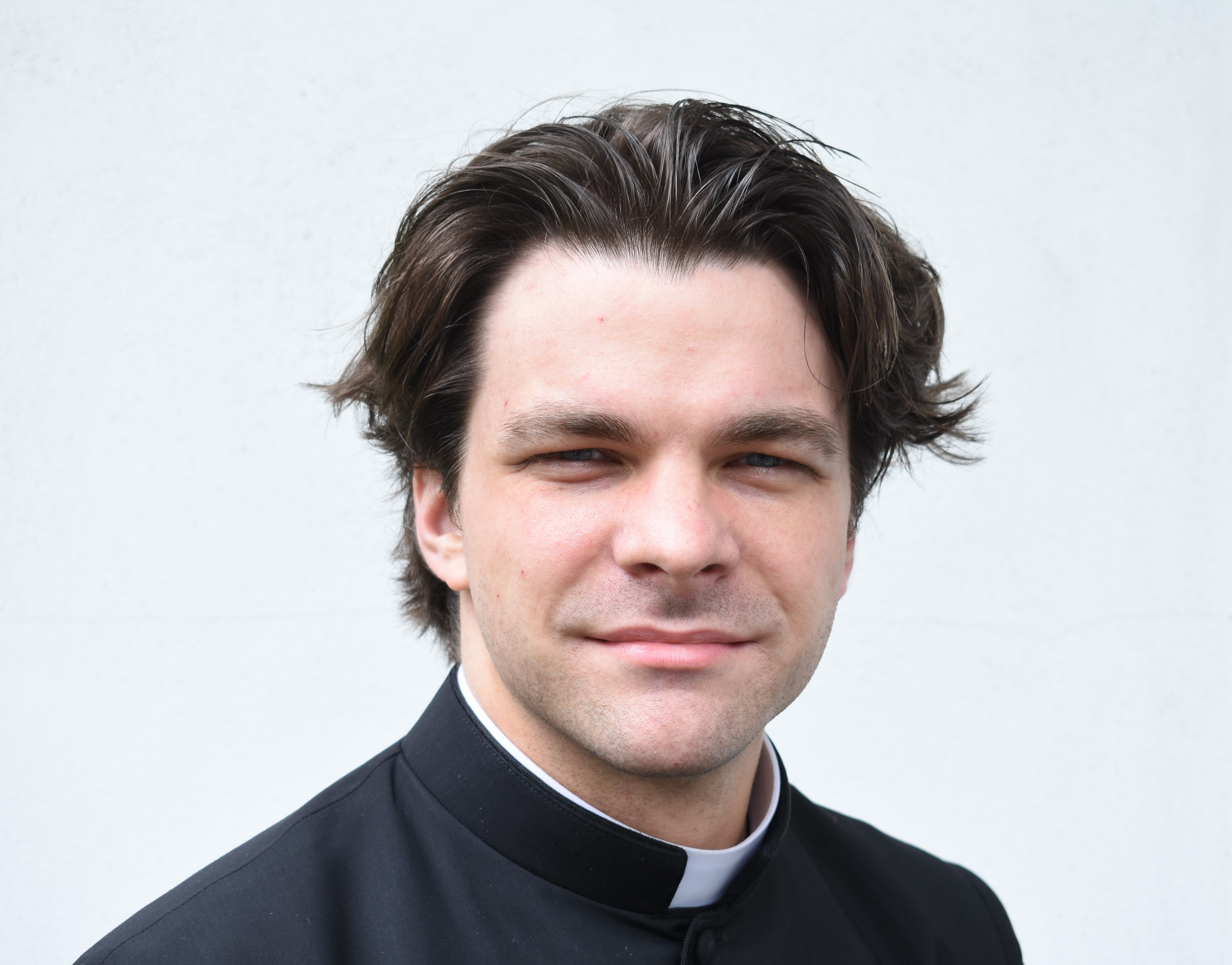 A white man with brown hair wears a clerical collar. His hair is shorter on the sides and longer on top.
