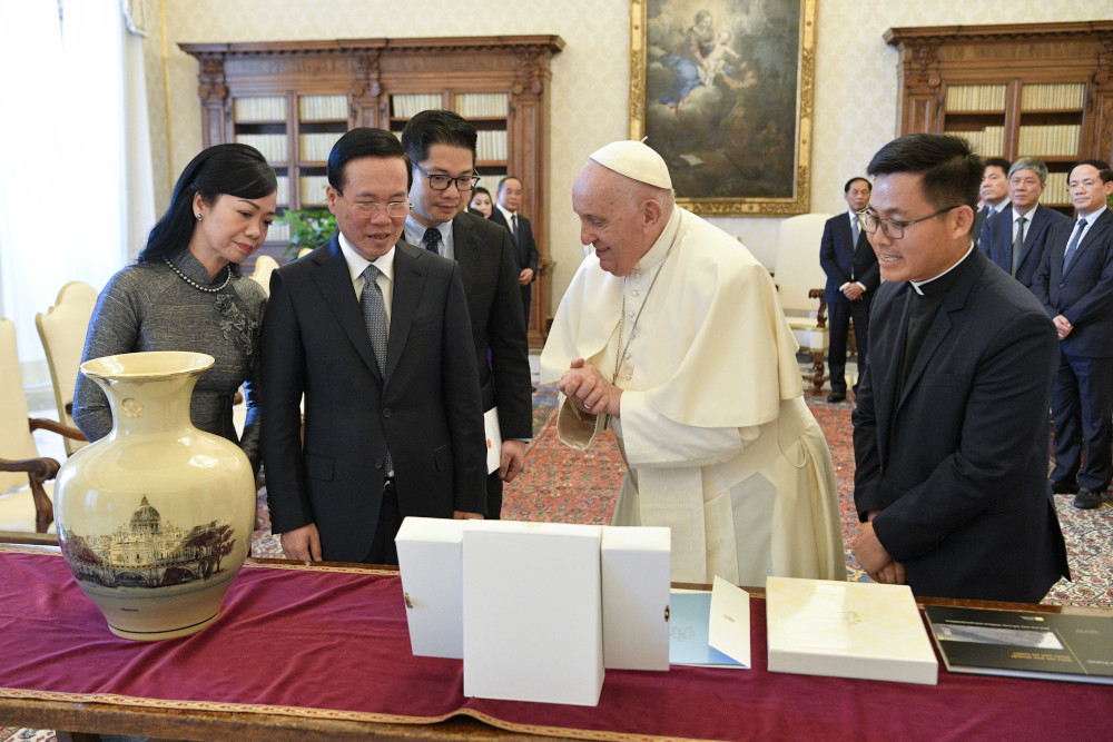 Pope Francis stands between a Vietnamese man in a suit and an Asian man in a clerical collar as they study objects on a table