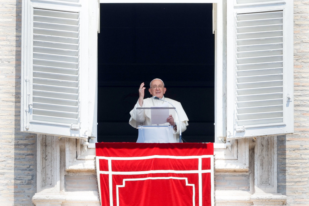Pope Francis stands at his apartment window and raises one hand as he speaks behind a clear lecturn
