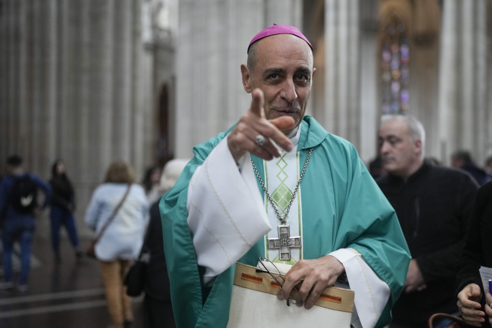 A man wearing green vestments and a violet zucchetto points at the camera while standing in a church